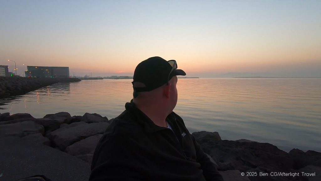 The author sitting in the area where the Sun Voyager monument is located in Reykjavik, Iceland. The ocean is calm and he is has his head turned away from the camera, looking at the colourful sky with the midnight sun, creating hues of blue, lavender and orange.